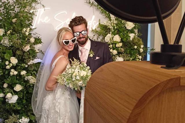 Una coppia di sposi posa con occhiali divertenti davanti alla postazione Photo Booth in legno