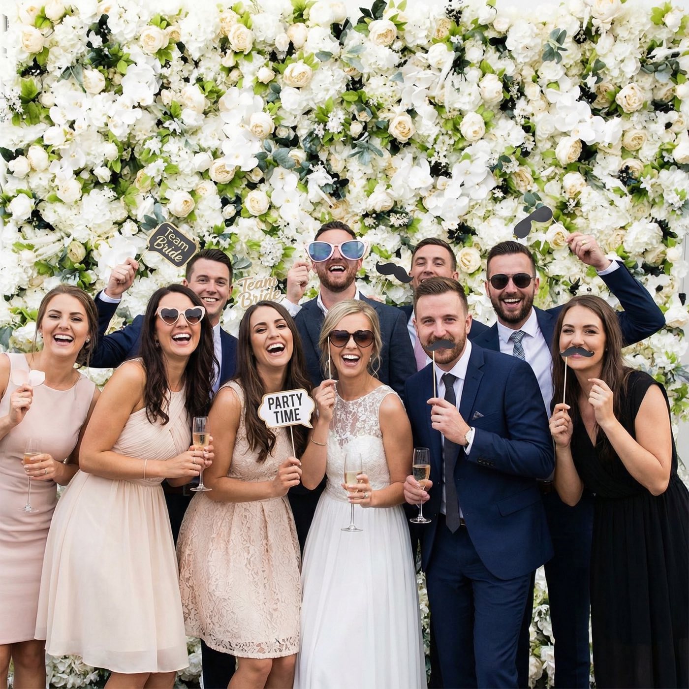Sposi e amici ridono insieme davanti a una parete di fiori, con occhiali e accessori Photo Booth